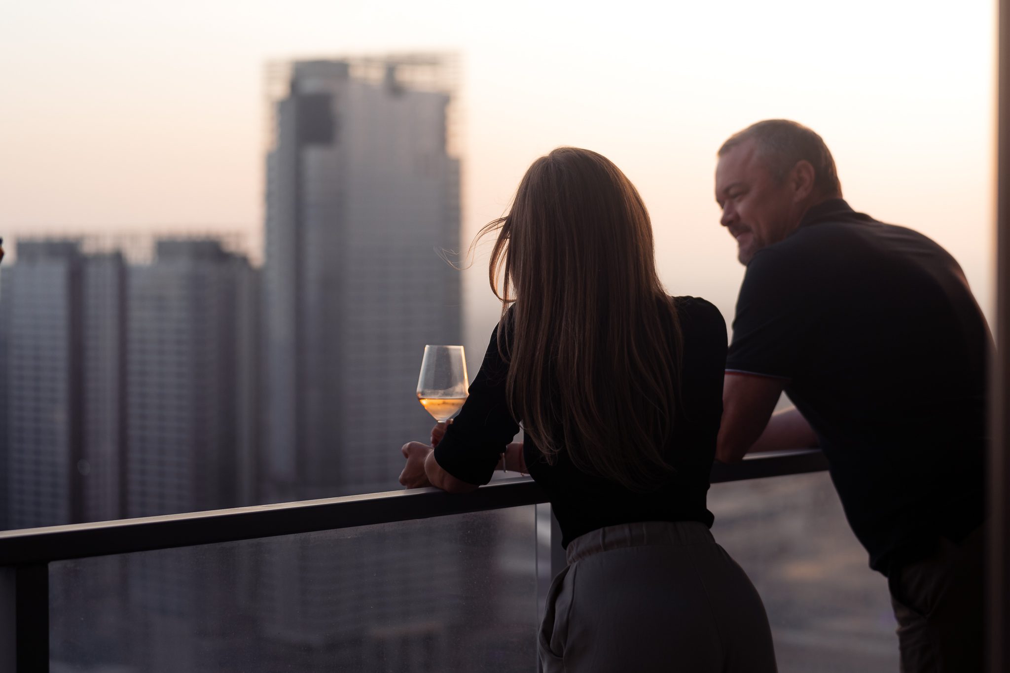 A man and a woman stand on a balcony at sunset, overlooking city skyscrapers. The woman holds a glass of wine as they relax—perhaps making the most of third night free hotel stays in Chicago, Los Angeles, or Seattle.