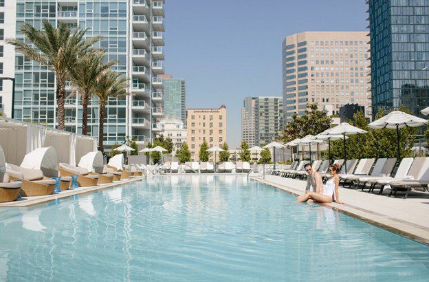 Two women sit at the edge of a clear swimming pool surrounded by lounge chairs and umbrellas, with tall modern buildings and palm trees in the background under a clear blue sky.