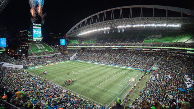 A large, packed soccer stadium at night with fans in the stands, players on the field, and fireworks going off above the scoreboard. The arena is brightly lit and energetic.