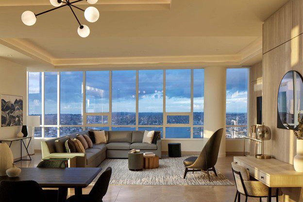 A modern living room with large windows offering a city and water view, featuring a gray sectional sofa, contemporary chairs, a rug, and stylish lighting in a warm, spacious setting.