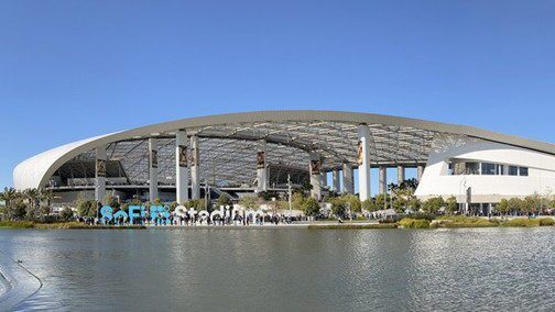 SoFi Stadium with its distinctive curved roof and open sides, reflected in a large pond under a clear blue sky, with people gathered near the entrance.