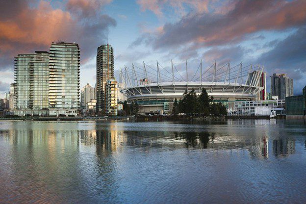 A stadium with a unique spiked roof is surrounded by modern high-rise buildings, all reflecting in the calm water under a dramatic, cloudy sky at sunset.