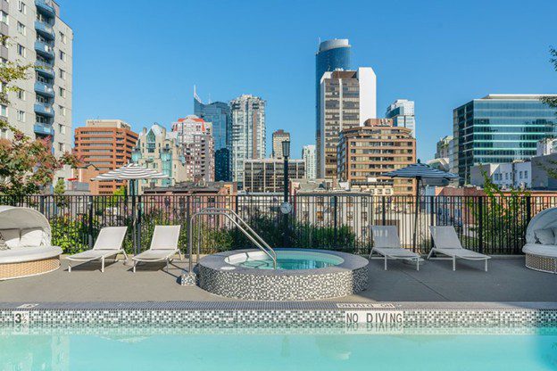 A rooftop pool and hot tub with lounge chairs overlooks a city skyline featuring modern high-rise buildings, under a clear blue sky.