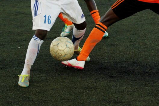 Two soccer players compete for the ball on a dark field, evoking the intensity of World Cup soccer in Vancouver. One wears a white uniform with blue accents and number 16; the other sports orange and black. Only their legs and the ball are visible.