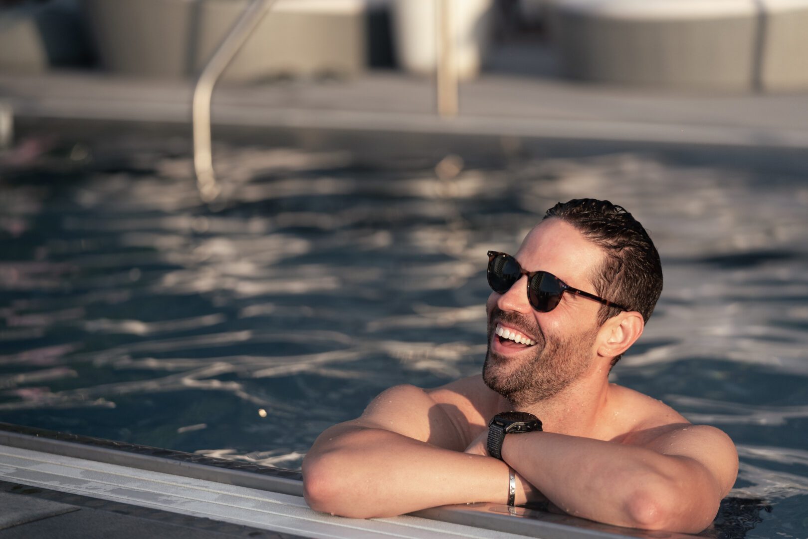 A man wearing sunglasses smiles and relaxes at the edge of a swimming pool, leaning his arms on the poolside as sunlight reflects off the water.
