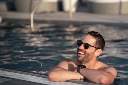 A man wearing sunglasses smiles and relaxes at the edge of a swimming pool, leaning his arms on the poolside as sunlight reflects off the water.