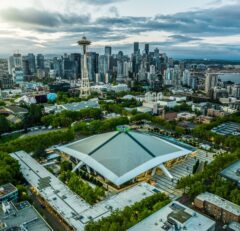 Aerial view of downtown Seattle with the Space Needle and Climate Pledge Arena surrounded by trees and city buildings under a partly cloudy sky.