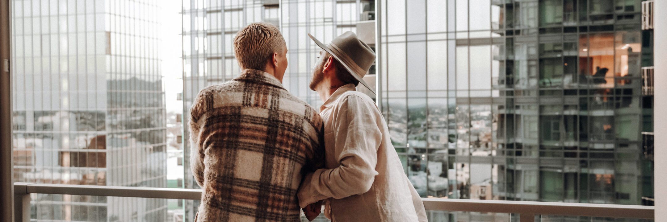 Two people stand close together on a balcony, looking out at a cityscape with tall glass buildings. One person wears a plaid jacket, and the other wears a wide-brimmed hat and light-colored clothing.