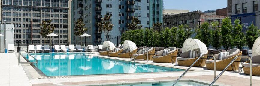 A modern rooftop pool area with lounge chairs, covered daybeds, and a small hot tub, surrounded by city buildings and greenery under a clear blue sky.